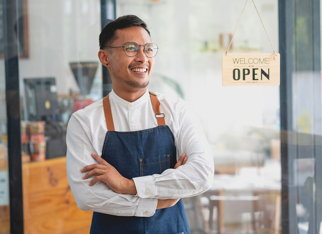 Business Insurance - Portrait of a Cheerful Middle Aged Small Business Owner Wearing an Apron Standing in Front of the Entrance of his Coffee Shop with his Arms Folded