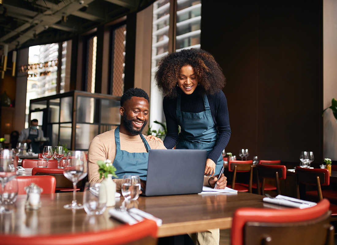 Insurance by Industry - Portrait of Two Cheerful Small Business Owners Working on a Laptop While Sitting at a Table Inside Their Restaurant